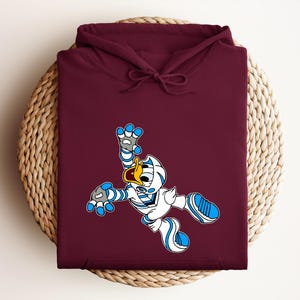 Peut inclure: Un sweat &agrave; capuche bordeaux avec un personnage de dessin anim&eacute; en tenue blanche et bleue. Le personnage a un bec jaune et est dans une pose dynamique. Le sweat &agrave; capuche est pli&eacute; sur une surface circulaire tiss&eacute;e.