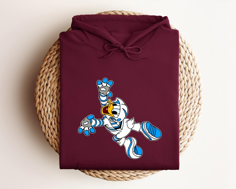 Peut inclure: Un sweat &agrave; capuche bordeaux avec un personnage de dessin anim&eacute; en tenue blanche et bleue. Le personnage a un bec jaune et est dans une pose dynamique. Le sweat &agrave; capuche est pli&eacute; sur une surface circulaire tiss&eacute;e.