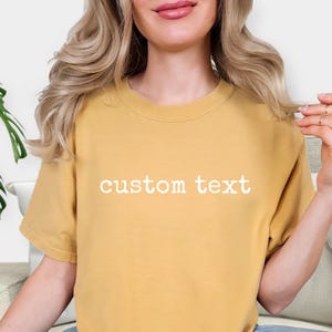 May include: A mustard yellow short-sleeved t-shirt featuring the words custom text printed in white across the center of the chest. The garment has a crew neckline and a relaxed fit, showcasing a solid color design with clear white lettering.