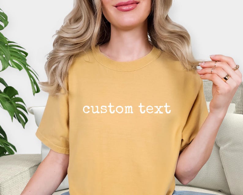May include: A mustard yellow short-sleeved t-shirt featuring the words custom text printed in white across the center of the chest. The garment has a crew neckline and a relaxed fit, showcasing a solid color design with clear white lettering.
