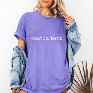 May include: A purple short-sleeved t-shirt featuring the words custom text printed in white across the chest. The shirt has a crew neckline and a relaxed fit, worn with a light blue denim jacket draped over the shoulders.