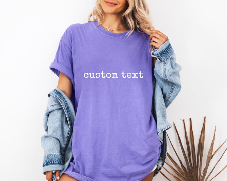 May include: A purple short-sleeved t-shirt featuring the words custom text printed in white across the chest. The shirt has a crew neckline and a relaxed fit, worn with a light blue denim jacket draped over the shoulders.