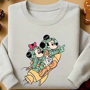 Peut inclure: Un sweat-shirt gris clair avec une illustration de dessin anim&eacute; de Mickey et Minnie Mouse en combinaisons spatiales sur une fus&eacute;e jaune. Les noms "Amelia" et "Logan" sont sur les casques. Le design comprend des accents verts, roses et oranges.