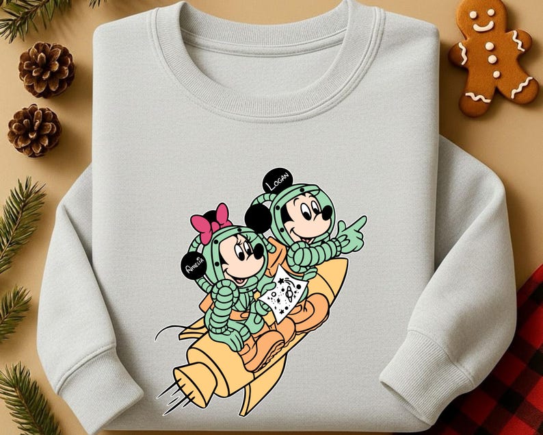 Peut inclure: Un sweat-shirt gris clair avec une illustration de dessin anim&eacute; de Mickey et Minnie Mouse en combinaisons spatiales sur une fus&eacute;e jaune. Les noms "Amelia" et "Logan" sont sur les casques. Le design comprend des accents verts, roses et oranges.