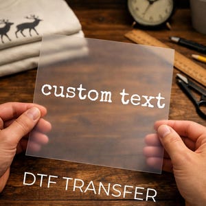 May include: A square, semi-transparent sheet featuring the white text custom text printed in a typewriter-style font. This DTF transfer is held up against a dark wooden surface, with a stack of folded shirts and various office supplies visible in the background.