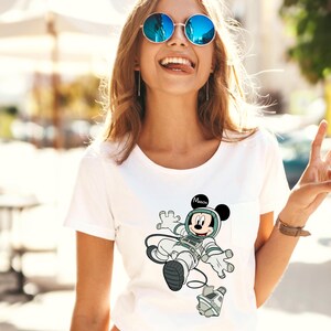 Peut inclure: T-shirt blanc avec un dessin de Mickey Mouse en astronaute. Le design comprend un casque et une combinaison spatiale gris et blancs, avec le mot "Mission" au-dessus de sa t&ecirc;te.
