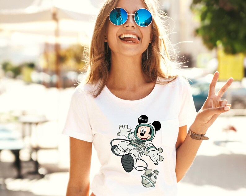 Peut inclure: T-shirt blanc avec un dessin de Mickey Mouse en astronaute. Le design comprend un casque et une combinaison spatiale gris et blancs, avec le mot "Mission" au-dessus de sa t&ecirc;te.