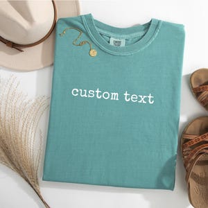 May include: A teal short-sleeved t-shirt featuring the white text custom text printed on the front. The shirt is displayed alongside a beige hat with a brown band, a gold-toned necklace, and a pair of brown strappy sandals with patterned details.