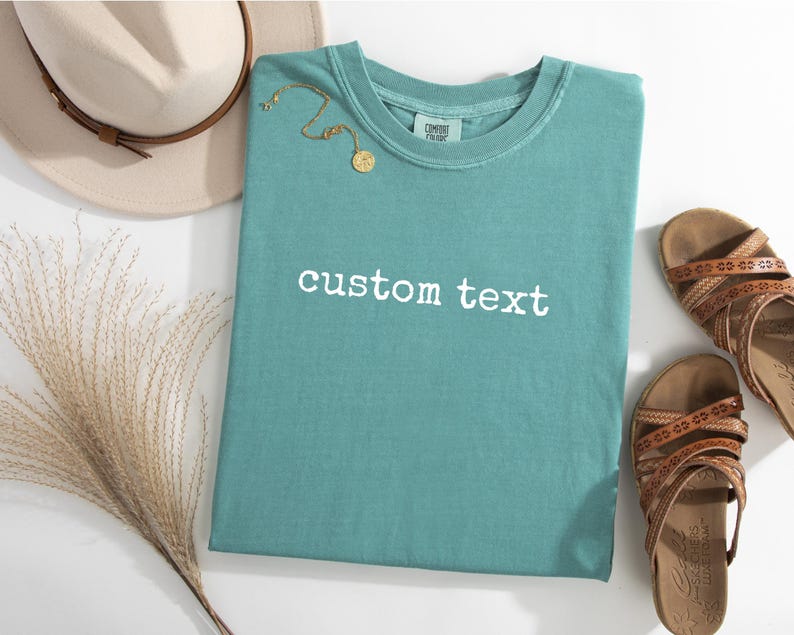 May include: A teal short-sleeved t-shirt featuring the white text custom text printed on the front. The shirt is displayed alongside a beige hat with a brown band, a gold-toned necklace, and a pair of brown strappy sandals with patterned details.
