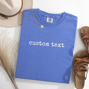 May include: A periwinkle blue short-sleeve t-shirt featuring the white text custom text printed on the front. The shirt has a crew neckline and a Comfort Colors brand label on the inside collar. A gold chain necklace rests on the fabric.