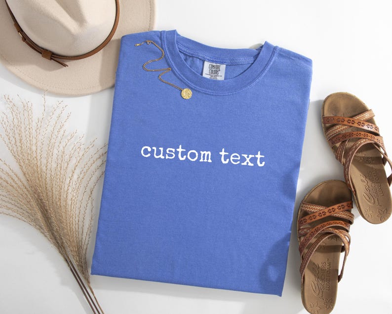 May include: A periwinkle blue short-sleeve t-shirt featuring the white text custom text printed on the front. The shirt has a crew neckline and a Comfort Colors brand label on the inside collar. A gold chain necklace rests on the fabric.