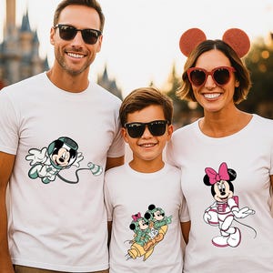 Peut inclure: Trois personnes portant des t-shirts blancs avec des motifs de personnages de dessins anim&eacute;s. Les chemises pr&eacute;sentent Mickey et Minnie Mouse en tenues d'astronaute. La famille sourit et porte des lunettes de soleil, posant &agrave; l'ext&eacute;rieur.