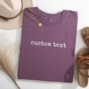 May include: A plum-colored short-sleeve t-shirt featuring the white embroidered text custom text centered on the chest. The shirt is displayed alongside a beige fedora hat, a gold-toned necklace with a circular pendant, and two brown sandals.