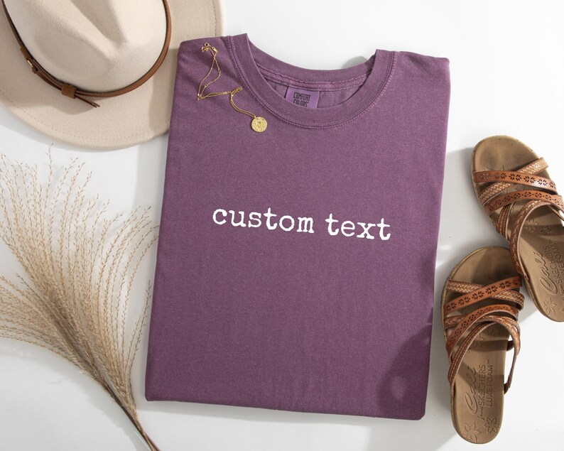 May include: A plum-colored short-sleeve t-shirt featuring the white embroidered text custom text centered on the chest. The shirt is displayed alongside a beige fedora hat, a gold-toned necklace with a circular pendant, and two brown sandals.
