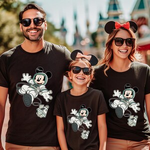 Peut inclure: Trois personnes portant des t-shirts noirs assortis avec un dessin anim&eacute; de Mickey Mouse en combinaison spatiale. Les chemises portent les noms "Mason" et "Emilia". La famille sourit et porte des lunettes de soleil.