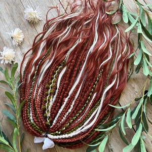 May include: A set of synthetic hair extensions featuring a mix of auburn, olive green, and white strands. The collection includes a variety of textures, such as tight braids, loose wavy locks, and thin yarn-like accents, all gathered at one end.