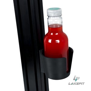Peut inclure: Un porte-gobelet en plastique noir fixé sur un profilé d'extrusion en aluminium noir vertical. Une bouteille en plastique transparent remplie d'un liquide rouge vif avec un bouchon bleu clair est placée dedans. Le logo LAKE PIT est visible.