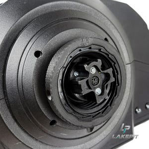 以下が含まれることがあります： 黒い円形のレーシングステアリングホイールハブアダプター。テクスチャ加工された外輪、中央のマルチピン電気コネクタ、および目に見えるネジを備えた十字型の取り付けプレートが特徴です。右下にLAKEPIのロゴが印刷されています。