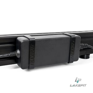 May include: A black rectangular power adapter brick secured to a black metal rail by two black rubber straps. The word LAKEPIT is embossed on both straps. A black power cable extends from the left side of the adapter, and another cable extends from the right.