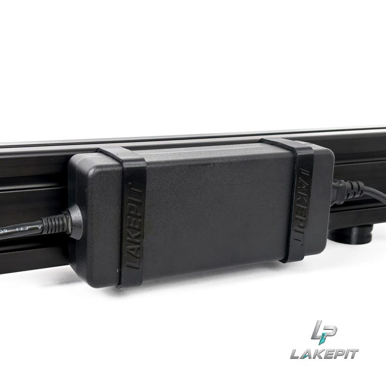 May include: A black rectangular power adapter brick secured to a black metal rail by two black rubber straps. The word LAKEPIT is embossed on both straps. A black power cable extends from the left side of the adapter, and another cable extends from the right.