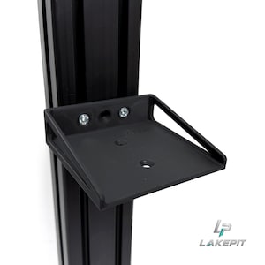 Puede incluir: Un soporte de montaje negro fijado a un perfil de extrusión de aluminio negro vertical. El soporte cuenta con una plataforma plana y rectangular con soportes laterales y dos tornillos plateados en la parte trasera. El texto LAKE PIT está grabado.