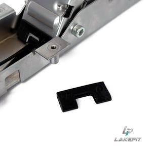 Könnte beinhalten: Eine silberfarbene mechanische Scharnierkomponente aus Metall mit einem Sechskantschraubenkopf und einer kleinen, flachen, rechteckigen Metallplatte mit einem Gewindeloch. Ein separates, kleines, schwarzes U-förmiges Kunststoff-Distanzstück liegt daneben.
