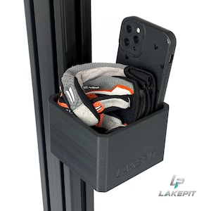 Op de afbeelding: Een zwarte rechthoekige opbergbak met de tekst LAKEPIT in reliëf op de voorzijde, gemonteerd op een verticale zwarte aluminium extrusierail. In de bak liggen een zwarte smartphone met een hartjespatroon en een paar grijze, zwarte en oranje handschoenen.