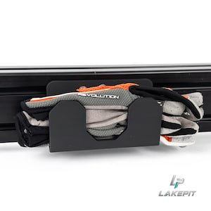 Op de afbeelding: Een zwarte metalen opberghouder gemonteerd op een horizontale zwarte rail, met daarin een paar grijze, zwarte en oranje handschoenen. De handschoenen hebben het woord REVOLUTION in het wit op de bovenkant en de houder heeft een uitsparing.