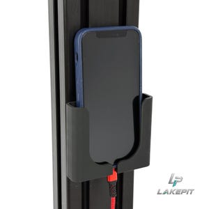 May include: A black plastic smartphone wall mount attached to a vertical black aluminium extrusion profile. A smartphone with a dark blue case is resting inside the mount, connected to a red charging cable at the bottom. The logo LAKE PIT is visible.