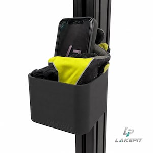 Puede incluir: Un contenedor de almacenamiento negro mate con el texto LAKEPIT en relieve, fijado a un perfil de extrusión de aluminio negro vertical. Dentro del contenedor hay un teléfono inteligente negro y un par de guantes negros y amarillo neón.