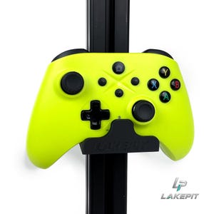 Könnte beinhalten: Ein neongelber Videospiel-Controller, der auf einer schwarzen Wandhalterung montiert ist, auf der das Wort LAKEPIT eingeprägt ist. Der Controller verfügt über schwarze Analogsticks, ein schwarzes Steuerkreuz und vier farbige Aktionstasten mit X, Y, A und B.
