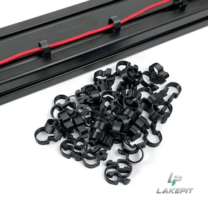 Op de afbeelding: Een verzameling kleine zwarte C-vormige kabelclips gerangschikt in een hoop, vergezeld van een lange zwarte metalen rail met een rood koord vastgezet door twee identieke clips. Het logo LAKE PIT is zichtbaar in de rechterbenedenhoek.