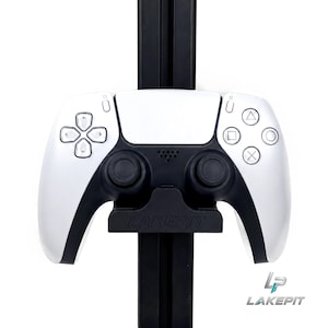 May include: A white and black wireless video game controller resting on a black mounting bracket. The controller features a directional pad, two analog sticks, and four action buttons with geometric symbols. The word LAKEPIT is embossed on the mount.