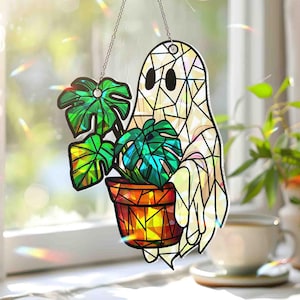 May include: A stained glass ghost holding a potted plant, suspended by a chain. The ghost is white with black eyes and a geometric design. The plant has green leaves and sits in an orange and yellow pot. Text at the bottom reads "Broken Glass Hologram."