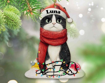 May include: A black and white cat wearing a red Santa hat with the name "Luna" on it. The cat is surrounded by colorful Christmas lights and a red ornament. The cat is sitting on a white snowy surface.