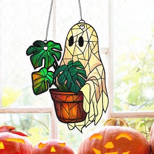 May include: A stained glass ghost holding a potted Monstera plant, suspended by a chain. The ghost is off-white with black eyes and a geometric design. The plant has green leaves and sits in a brown pot. Carved pumpkins are in the foreground.