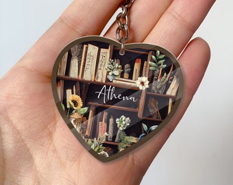 May include: A heart-shaped keychain with a brown wooden bookshelf design. The bookshelf is filled with books, plants, and candles. The text "Athena" is written in white cursive on the front of the keychain.