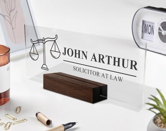 May include: A clear acrylic desk sign with a black scales of justice icon and the text "JOHN ARTHUR SOLICITOR AT LAW". The sign is on a wooden stand.