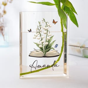 May include: Clear acrylic vase with a floral and butterfly design. The vase features a lily of the valley illustration over an open book, with the name "Amanda" in script. Green stems and leaves are visible inside the vase.
