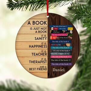 May include: A round wooden ornament with a quote about books. The quote reads "A book is just not a book. It is sanity, it is happiness, it is teacher, it is therapist, and a best friend." The ornament is decorated with a stack of books with titles on the spines. The name "Daniel" is written at the bottom of the ornament.