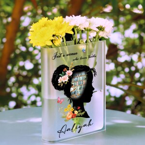 May include: A clear glass vase with a silhouette of a woman reading a book. The vase has the text "Just a woman who loves books" and the name "Aaliyah" printed on it. The vase is filled with yellow and white flowers.