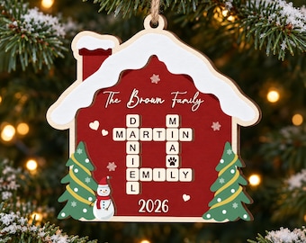 May include: A house-shaped wooden Christmas ornament in red with white snow on the roof. It features a crossword-style grid with the names Daniel, Martin, Mia, and Emily, the text The Brown Family, a snowman, two green trees, and the year 2026.
