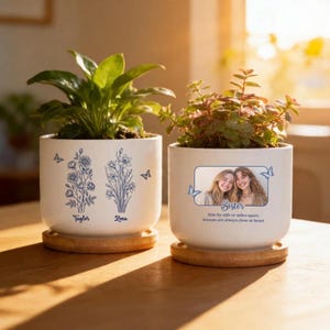 May include: Two white ceramic planters with plants, each on a wooden coaster. One planter has blue floral designs and the names "Taylor" and "Zena." The other features a photo and the text "Sister." Blue butterflies decorate both planters.