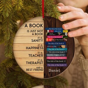 May include: A wooden Christmas ornament shaped like a book with the text "A book is just not a book. It is sanity. It is happiness. It is teacher. It is therapist. And a best friend."  The ornament is decorated with a wood grain pattern and has a red ribbon attached to the top. The back of the ornament is decorated with a stack of books with the titles "The title of the book 1", "The title of the book 2", "The title of the book 3", "The title of the book 4", "The title of the book 5", "The title of the book 6", "The title of the book 7", "The title of the book 8", "The title of the book 9", "The title of the book 10", "The title of the book 11", "The title of the book 12", "The title of the book 13". The name "Daniel" is written at the bottom of the ornament.