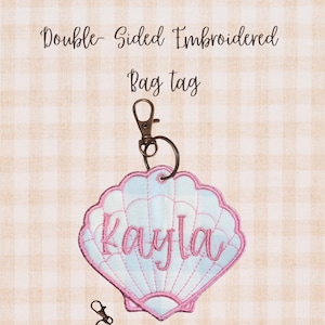May include: Double-sided embroidered bag tag in a seashell shape. The tag is light blue with pink trim and the name "Kayla" embroidered in pink. Includes a smaller matching tag and a bronze-coloured clasp.