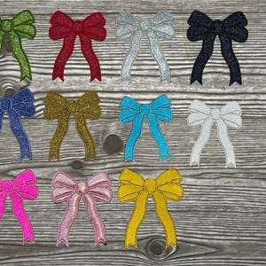 May include: Twelve different coloured glitter bows with a variety of colours including red, blue, green, gold, silver, pink, and turquoise. The bows are arranged in two rows of six on a wooden background.