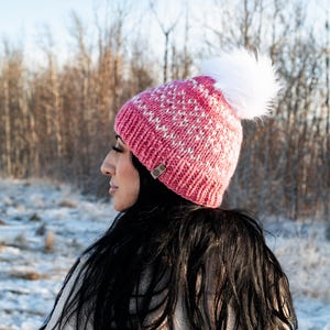 May include: A pink and white knit beanie with a white faux fur pom pom. The beanie has a snowflake pattern.