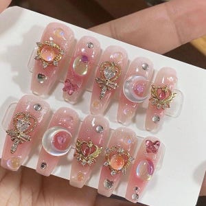 Peut inclure: Faux ongles longs roses et blancs avec des accents de paillettes dorées et argentées. Chaque ongle est décoré d'un charme différent, y compris un croissant de lune, un cœur, une étoile et un papillon.