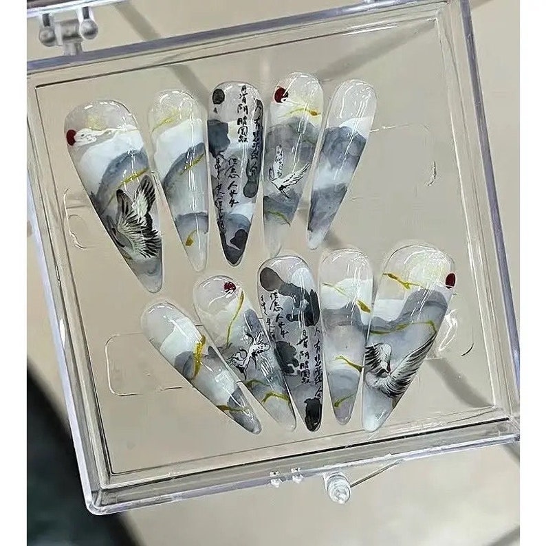 May include: A set of ten false fingernails with a white, gray, and gold design. The nails feature a crane bird, clouds, and Japanese calligraphy.