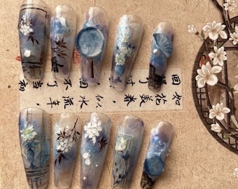 May include: Ten long, clear, acrylic false nails with blue and white floral designs. The designs include flowers, branches, and a fan. The nails are arranged in two rows on a beige surface with Chinese calligraphy.